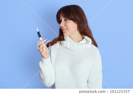 Young attractive female with pleasant appearance, lady wearing white sweater standing against blue wall and looking aside, girl with dark hair decides smoke or not. 101225357