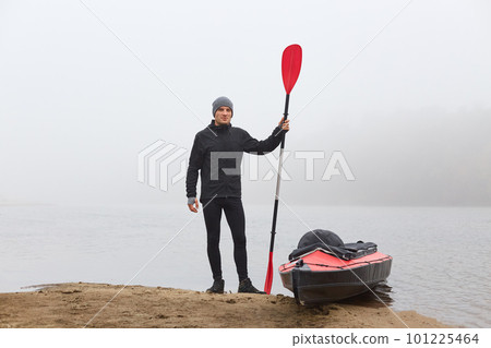 Young tourist in warm clothes stands on bank of river and assembles kayak to travel on icy river under grey sky Young tourist in warm clothes stands on bank of river and assembles kayak to travel on icy river under grey sky 101225464