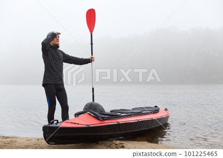 Full length shot of handsome young man wearing black clothing and cap, holding oar in hand and looking into distance with palm near forehead, sports man planning his way canoeing. Full length shot of handsome young man wearing black clothing and cap, holding oar in hand and looking into distance with palm near forehead, sports man planning his way canoeing. 101225465