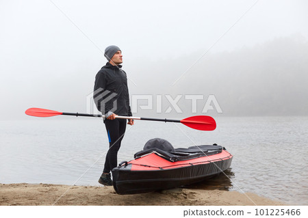 Young man athlete carrying canoe oar, looking into distance, wearing black trousers, jacket and gray cap, holding red oar in hand, looking into distance with dreamily expression. 101225466