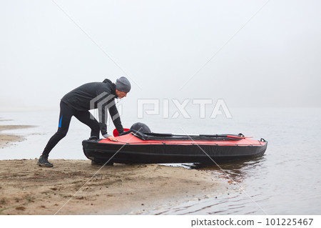 Man pulling canoe into water, wearing black clothing, posing on bank of river, handsome sports man doing water sport in foggy autumn day, canoeing in cloudy morning. 101225467