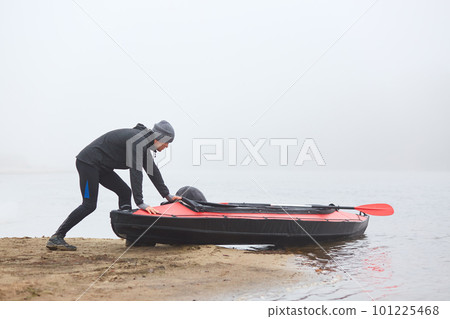 Young sportsman being ready rowing canoe, pushing boat into water, kayaker in warm clothes, padding in early fogging morning, extreme water sport. Young sportsman being ready rowing canoe, pushing boat into water, kayaker in warm clothes, padding in early fogging morning, extreme water sport. 101225468