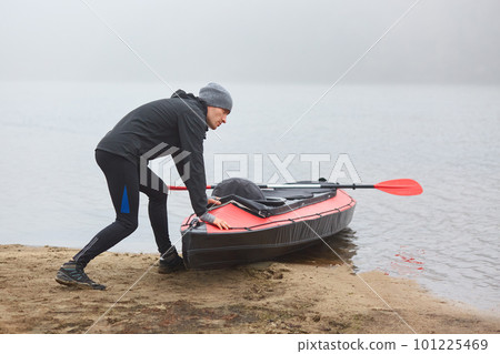 Lonely man going paddling on kayak in early foggy morning, sports man being fond of water sport, guy pushing boat into water, looks at misty river, wearing warm clothing. Lonely man going paddling on kayak in early foggy morning, sports man being fond of water sport, guy pushing boat into water, looks at misty river, wearing warm clothing. 101225469