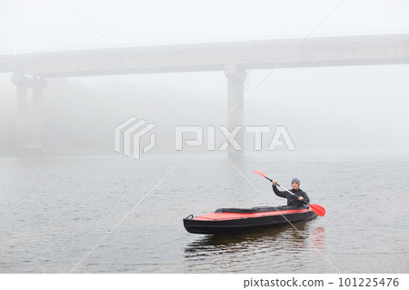Man canoeing in traditional wooden kayak on large lake at cold cloudy day, fog over water, handsome guy wearing black jacket and gray cap, kayaking alone in lake with bridge on backround. Man canoeing in traditional wooden kayak on large lake at cold cloudy day, fog over water, handsome guy wearing black jacket and gray cap, kayaking alone in lake with bridge on backround. 101225476