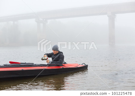 handsome young male in canoe in middle of river, pouring hot beverage from thermos, guy wearing black jacket and gray cap enjoying coffee or tea in kayak in open air on beautiful foggy autumn day. handsome young male in canoe in middle of river, pouring hot beverage from thermos, guy wearing black jacket and gray cap enjoying coffee or tea in kayak in open air on beautiful foggy autumn day. 101225478