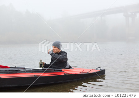 Side view of pensive guy in black jacket and gray cap, male sitting in canoe in middle of lake, spends his free time for his hobby, looks dreamily in distance, posing with bridge in fog on background. Side view of pensive guy in black jacket and gray cap, male sitting in canoe in middle of lake, spends his free time for his hobby, looks dreamily in distance, posing with bridge in fog on background. 101225479