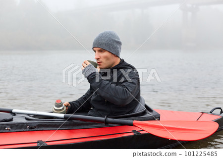Young attractive guy kayaking on river, stops in middle of river to have rest and have snack, holding thermos and drinks hot coffee or tea, man looking at water. 101225485