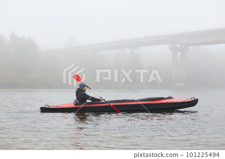 Side view of young kayaker in river, padding, man enjoying water sport in foggy morning with bridge on background, male rowing boat in cold autumn day. Side view of young kayaker in river, padding, man enjoying water sport in foggy morning with bridge on background, male rowing boat in cold autumn day. 101225494