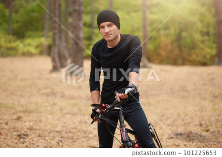 Mountain bike cyclist riding single track outdoor with trees on background, attractive male wearing black sport clothing and cap standing with his bicycle in forest, spending free time. Sport concept. Mountain bike cyclist riding single track outdoor with trees on background, attractive male wearing black sport clothing and cap standing with his bicycle in forest, spending free time. Sport concept. 101225653