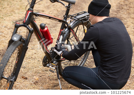 Outdoor shot of man repairing his icycle in field or forest, having puncture of bicycle camera on way, guy posing backwards, squating near his bike, trying to fix it, does sport while hawing free time 101225674