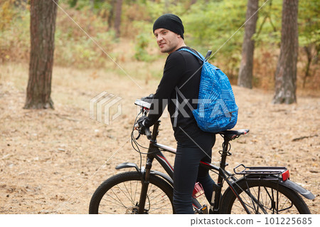 Young satisfied handsome man being in high spirits, riding his bicycle in forest alone, using GPS navigator on his smartphone, wearing black tracksuit, having blue backpack on his back. Sport concept. 101225685