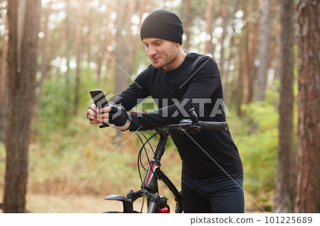 Horizontal outdoor shot of sportsman posing around nature with his bicycle, holding his mobile phone, touching its screen, using social networking sites, having short break. Technology concept. 101225689