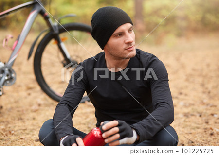 Picture of young man sitting near bike and wants to drink water from red bottle which holding in hands, looking thoughtfully aside, wearing black sportwear and cap, having recreation in forest. 101225725