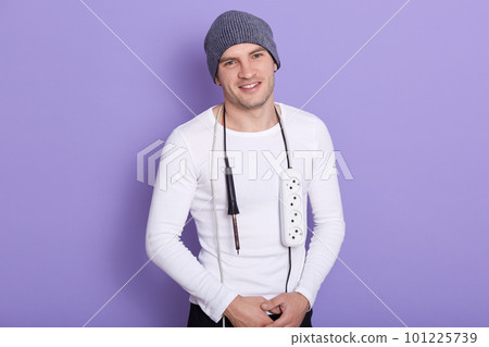 Indoor shot of young technician after soldering wire, attractive male wearing white casual shirt and gray cap, smiling male holding soldering iron around neck, posing isolated over purple background. 101225739