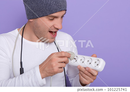 Shot of man tries to insert wire with electrical plug into main extension cord with sockets, young electrician with excited expression, wearing wite shirt and gray cap, isoalted over lilac background. Shot of man tries to insert wire with electrical plug into main extension cord with sockets, young electrician with excited expression, wearing wite shirt and gray cap, isoalted over lilac background. 101225741