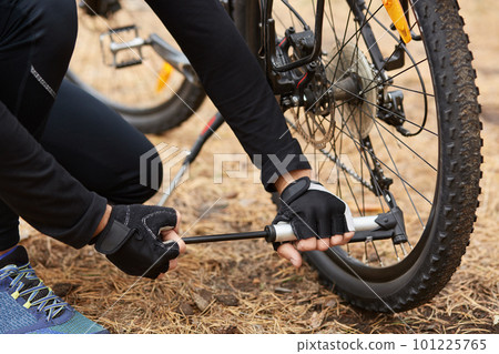 Cyclist in black sports wear holding pump in both hands, using it in pumping tires up, having problems with further movements, bicycle standing on forest road during repair. Bike repair concept. Cyclist in black sports wear holding pump in both hands, using it in pumping tires up, having problems with further movements, bicycle standing on forest road during repair. Bike repair concept. 101225765