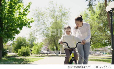 Happy family park. girl daughter rides bike for first time city park. mother teaches cheerful child girl ride bike. childhood dream travel. mother helps child girl ride bicycle by pedaling. traveling Happy family park. girl daughter rides bike for first time city park. mother teaches cheerful child girl ride bike. childhood dream travel. mother helps child girl ride bicycle by pedaling. traveling 101226599