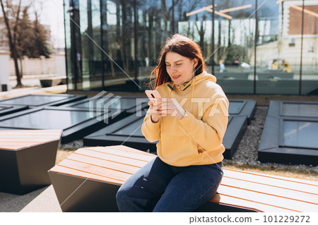 Young woman listening to music via headphones on the street. Pretty happy redhead woman standing on urban background and holding a mobile phone. Young woman listening to music via headphones on the street. Pretty happy redhead woman standing on urban background and holding a mobile phone. 101229272