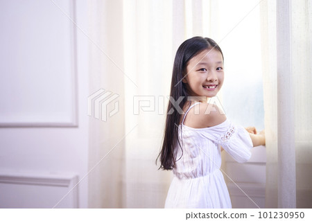 beautiful little asian girl with long black hair standing in front of windwo turning to camera smiling 101230950
