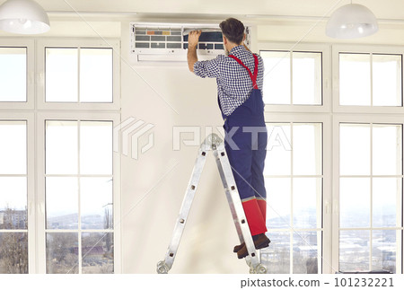 Professional electrician man standing back on a ladder maintaining modern air conditioner indoors. Professional electrician man standing back on a ladder maintaining modern air conditioner indoors. 101232221