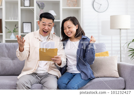Cheerful and happy asian family together, man and woman at home on sofa sitting in living room, received postal envelope with good news, message and good achievement news. Cheerful and happy asian family together, man and woman at home on sofa sitting in living room, received postal envelope with good news, message and good achievement news. 101232427