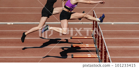 two female athletes running hurdles in athletics competition two female athletes running hurdles in athletics competition 101233415