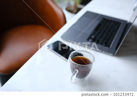 workplace with phone laptop and transparent cup of coffee in home office on white marble desk with brown leathern chair and green tree at background 101233472
