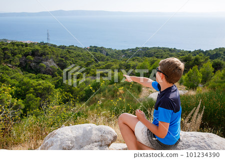 School boy sitting of hill in grass field and enjoying beautiful landscape view. Rear view of teenage boy hiker resting in nature. Active lifestyle. Concept of local travel School boy sitting of hill in grass field and enjoying beautiful landscape view. Rear view of teenage boy hiker resting in nature. Active lifestyle. Concept of local travel 101234390