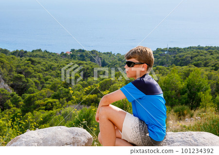 School boy sitting of hill in grass field and enjoying beautiful landscape view. Rear view of teenage boy hiker resting in nature. Active lifestyle. Concept of local travel School boy sitting of hill in grass field and enjoying beautiful landscape view. Rear view of teenage boy hiker resting in nature. Active lifestyle. Concept of local travel 101234392