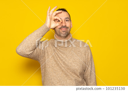 Handsome man over isolated yellow background doing ok gesture with hand smiling eye to eye through fingers with happy face Handsome man over isolated yellow background doing ok gesture with hand smiling eye to eye through fingers with happy face 101235130