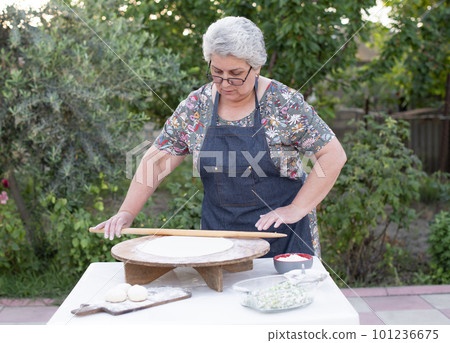 Front view of old female woman wearing apron looking down, rolling dough, making pita, taco outdoor. Concept of fast food. Front view of old female woman wearing apron looking down, rolling dough, making pita, taco outdoor. Concept of fast food. 101236675