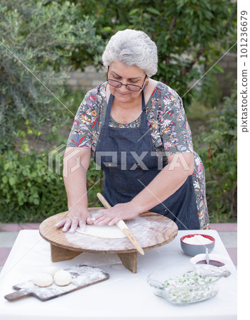 Front view of old female cook, chef wearing apron looking dowm , touching dough, rolling, making pita. Concept of fast food. 101236679