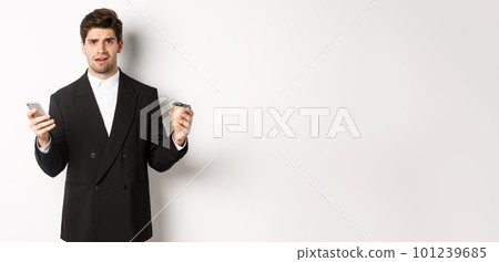 Image of confused businessman standing in a suit, holding cup of coffee and mobile phone, cant understand something, standing against white background 101239685