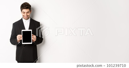 Image of good-looking male entrepreneur in black suit, looking down at digital tablet screen and showing advertisement, standing against white background 101239710