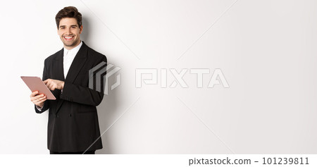 Image of handsome businessman in black suit, using digital tablet and smiling, standing against white background 101239811