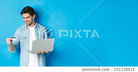 Young man order online, holding credit card and laptop, shopping in internet, standing against blue background Young man order online, holding credit card and laptop, shopping in internet, standing against blue background 101239815