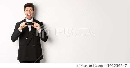Portrait of amazed handsome man in black suit, showing credit card and recommending bank, standing over white background 101239847