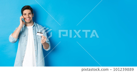 Handsome man calling bank and holding credit card, having mobile conversation, standing over blue background 101239889