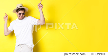 Tourism, travelling and holidays concept. Male tourist enjoying vacation, dancing in straw hat and sunglasses, pointing fingers sideways Tourism, travelling and holidays concept. Male tourist enjoying vacation, dancing in straw hat and sunglasses, pointing fingers sideways 101239901