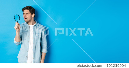 Young thoughtful guy looking through magnifying glass, reading something tiny, standing over blue background 101239904