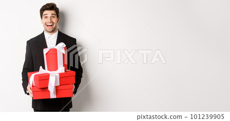 Concept of holidays, relationship and celebration. Handsome man in black suit bringing presents at new year party, holding gifts and smiling amused, standing against white background Concept of holidays, relationship and celebration. Handsome man in black suit bringing presents at new year party, holding gifts and smiling amused, standing against white background 101239905