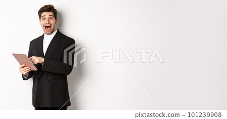 Portrait of amazed handsome businessman, wearing trendy suit, showing something cool on digital tablet, standing against white background 101239908