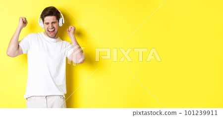 Happy man dancing and listening music in white headphones, standing over yellow background Happy man dancing and listening music in white headphones, standing over yellow background 101239911
