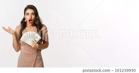 Shopping concept. Image of beautiful woman in glamour dress looking shocked, holding dollars and staring at camera, standing over white background 101239930