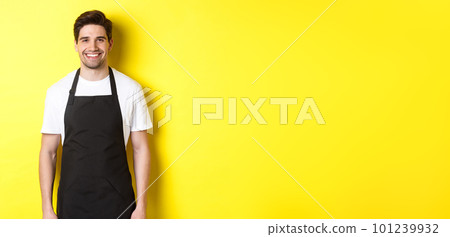 Young man waiter in black apron, smiling, working in store or coffee shop, standing against yellow background 101239932