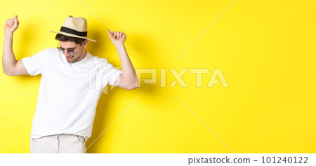 Tourism, travelling and holidays concept. Man tourist enjoying vacation, dancing in straw hat and sunglasses, posing against yellow background 101240122