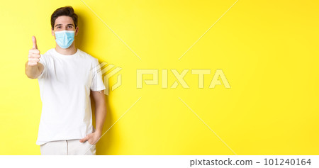 Concept of coronavirus, pandemic and social distancing. Confident young man in medical mask showing thumbs up, yellow background 101240164