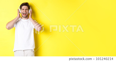 Happy guy listening music in new headphones, buying earphones on black friday, standing over yellow background 101240214