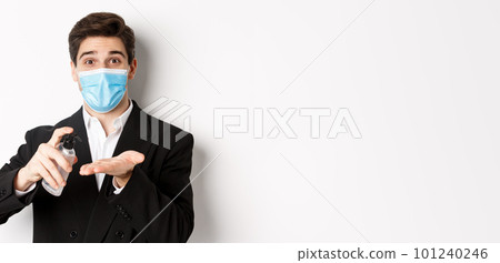 Concept of covid-19, business and social distancing. Image of handsome businessman in trendy suit and medical mask, cleaning hands with hand sanitizer, standing over white background Concept of covid-19, business and social distancing. Image of handsome businessman in trendy suit and medical mask, cleaning hands with hand sanitizer, standing over white background 101240246