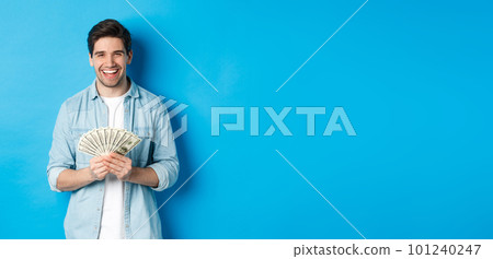 Happy successful man smiling pleased, holding money, standing over blue background Happy successful man smiling pleased, holding money, standing over blue background 101240247